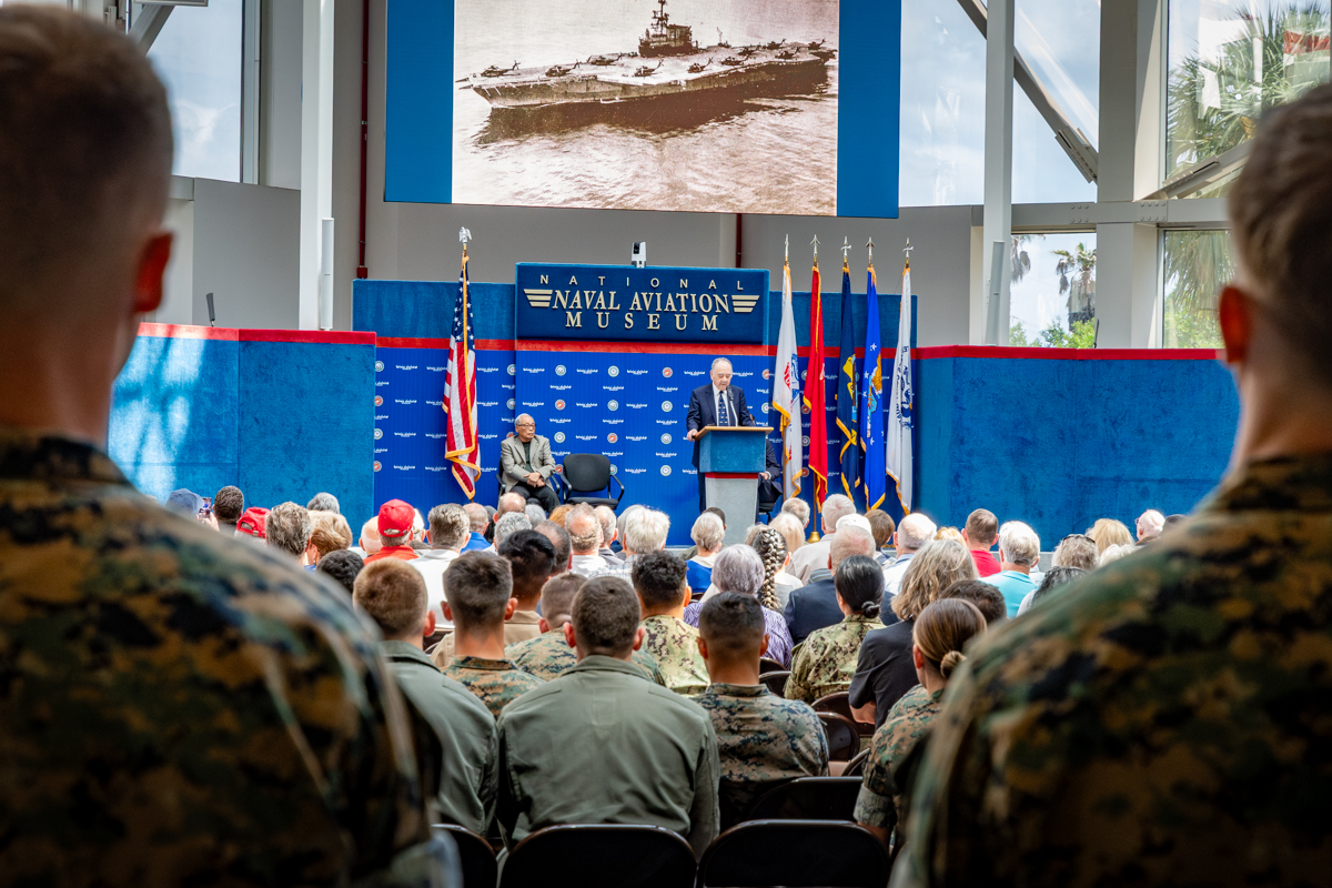 Speaker addresses audience at the National Naval Aviation Museum event, with military personnel and veterans in attendance, American flags and military branch flags on stage, and historic aircraft photo displayed above.