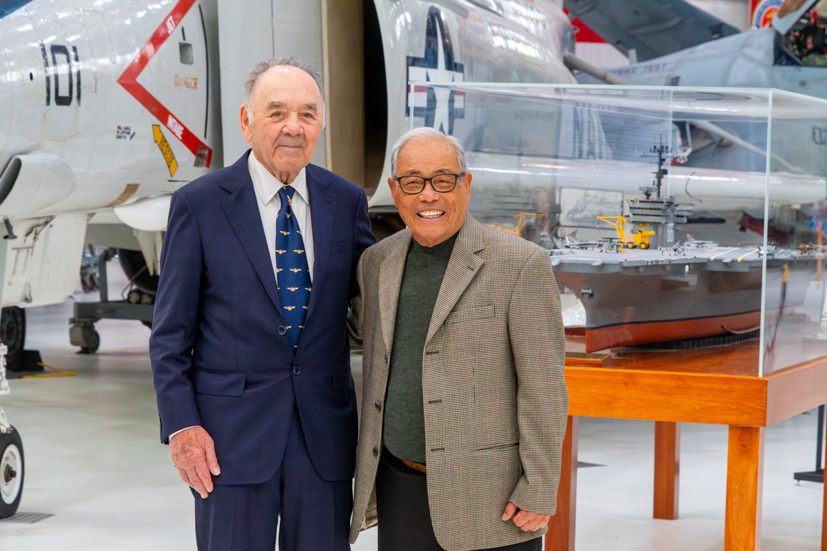 RADM Lawrence Chambers and Bung Lee pose smiling in front of a model aircraft carrier and a vintage Navy jet in Hangar Bay One at the National Naval Aviation Museum.