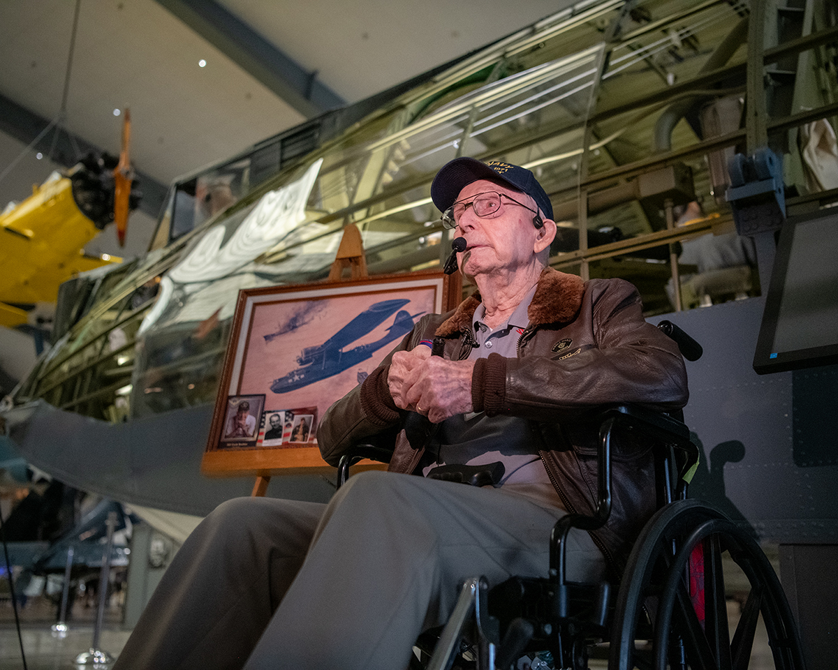 Cash Barber sitting in a wheel chair in front of an aircraft's cutaway display, telling his story to guests.