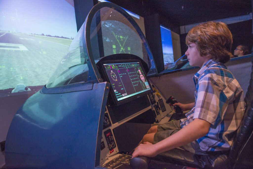 A young boy in a flight simulator cockpit at the National Naval Aviation Museum, focused on flying a virtual F-18 jet as part of a hands-on STEM activity during the Flight Adventure Deck Summer Camp.