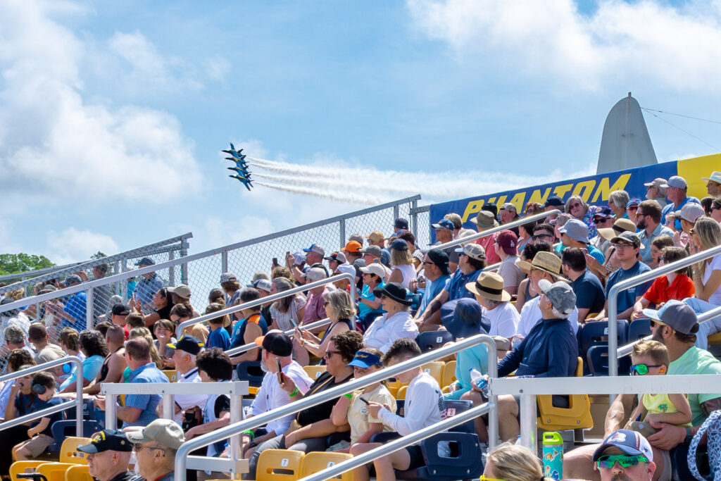 stadium bleachers filled with people watching the U.S. Navy Blue Angels flying in a bright, blue, cloudy sky.